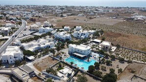 An aerial view of a coastal resort with white buildings, a central swimming pool, and a road curving through the property. - Perissa Hotel (Santorini)