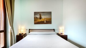 Ein ordentlich bezogenes Bett in einem Hotelzimmer, mit einem Gemälde an der Wand, zwei Nachttischlampen und einem Fenster mit Vorhängen.