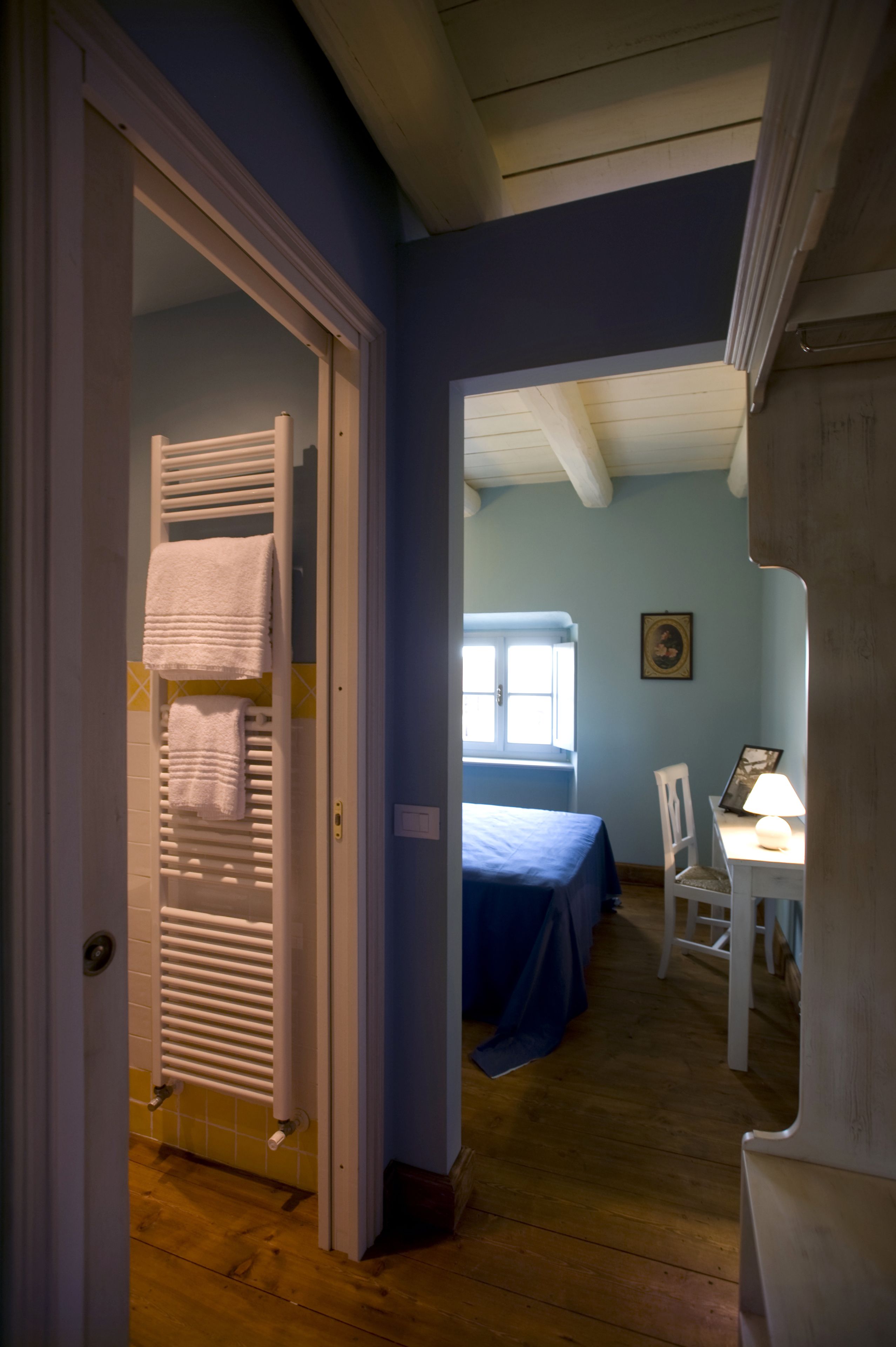 Een kamer met een bed, een bureau en een stoel. Een deur leidt naar een andere kamer met een radiator en handdoeken.