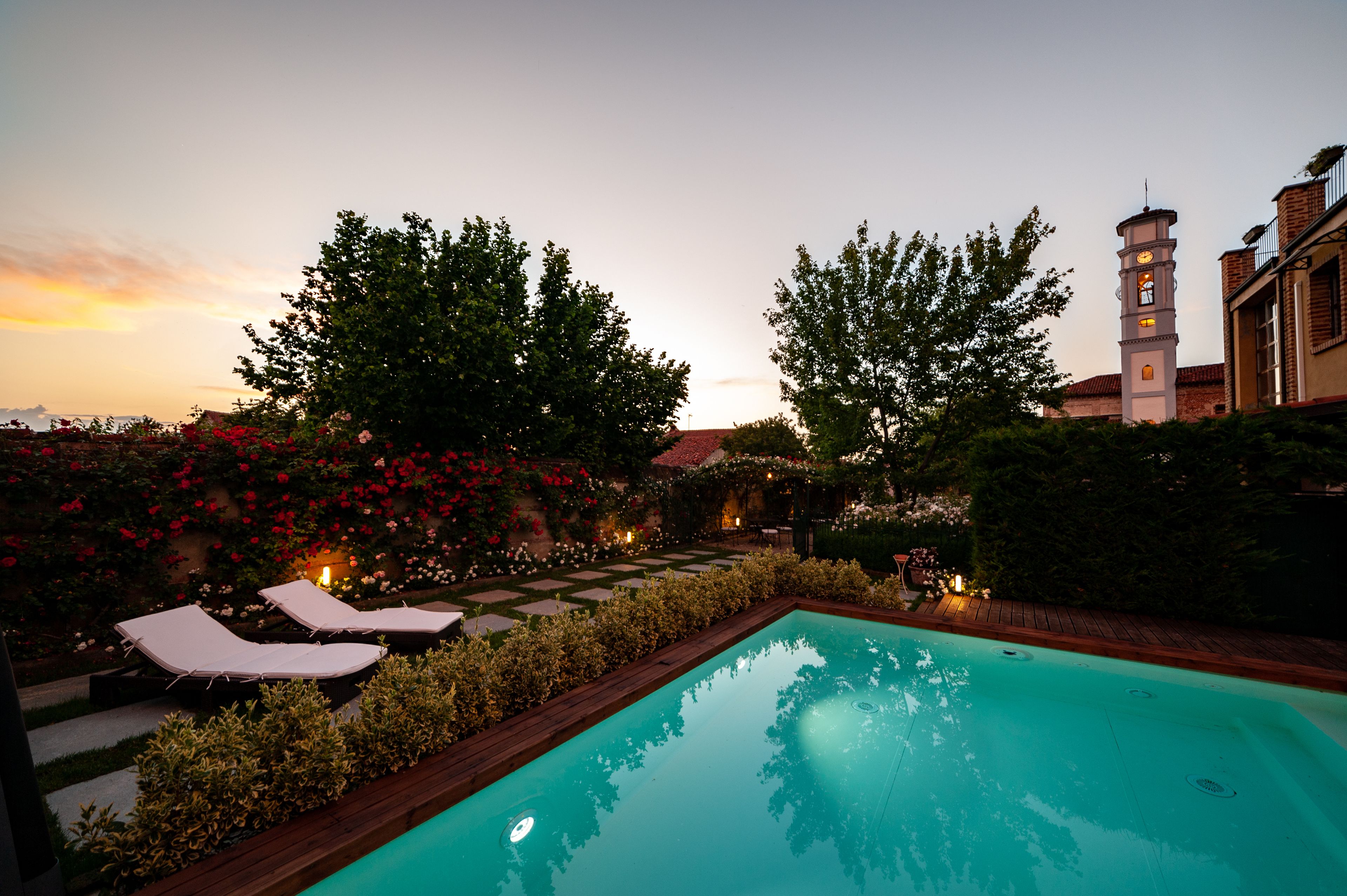 A swimming pool with lounge chairs, surrounded by a garden with flowering plants and a clock tower.
