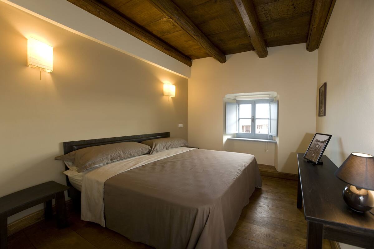 A bedroom with a bed, bedside lamps, a window, and a wooden ceiling.