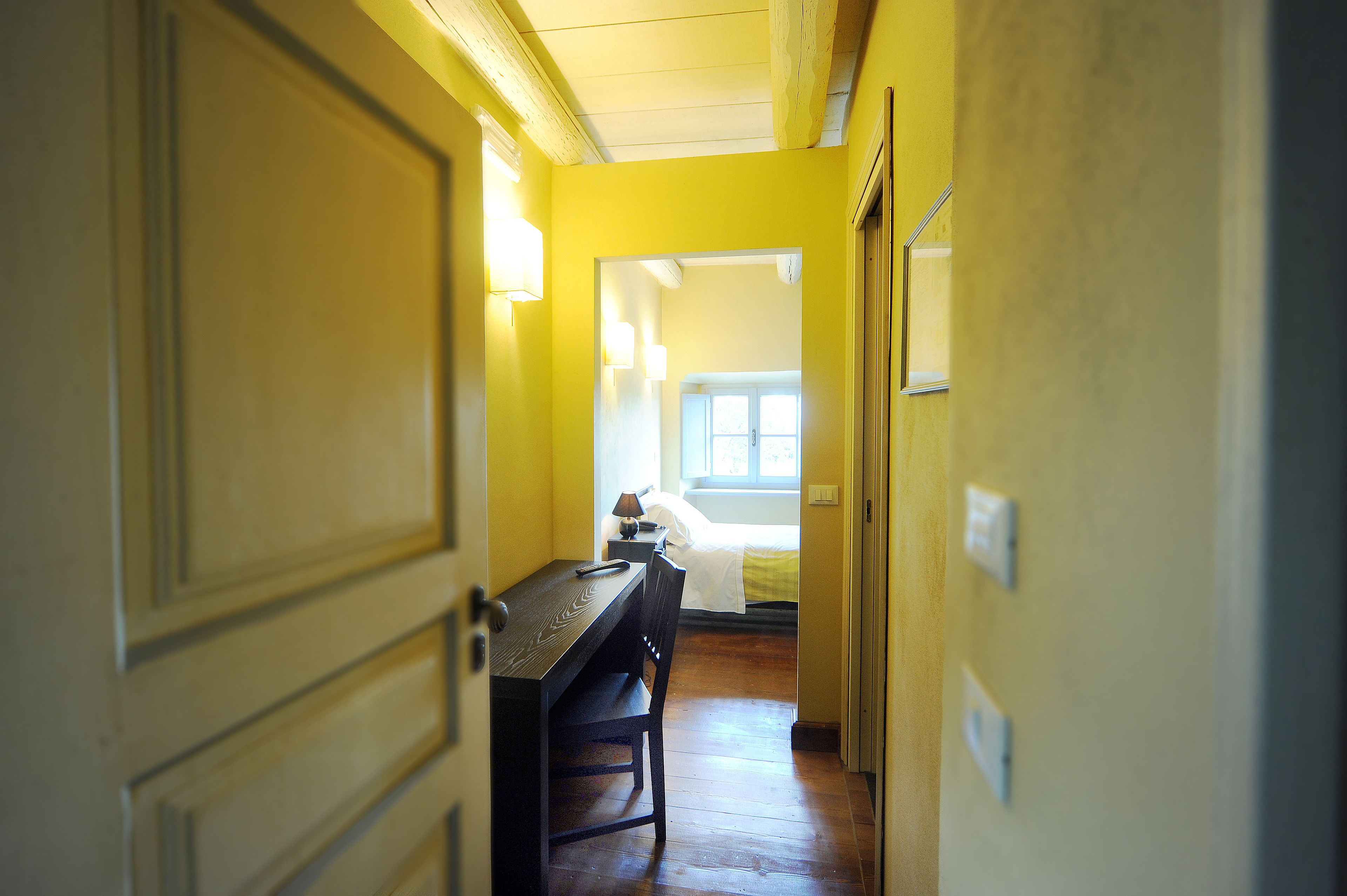 Un couloir avec un bureau et une chaise en bois, un lit recouvert d’une couette blanche et une fenêtre laissant entrer la lumière du jour.