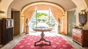 A hotel lobby with a central round table, a red patterned carpet, and a view of a garden through an arched doorway. - Hotel Il Campanile (Cherasco)