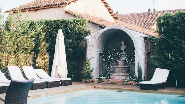 A pool area with lounge chairs, a fountain, and a building with a tiled roof. - Hotel Il Campanile (Cherasco)