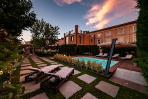 A pool area with lounge chairs, a stone pathway, and a brick building in the background. - Hotel Il Campanile (Cherasco)
