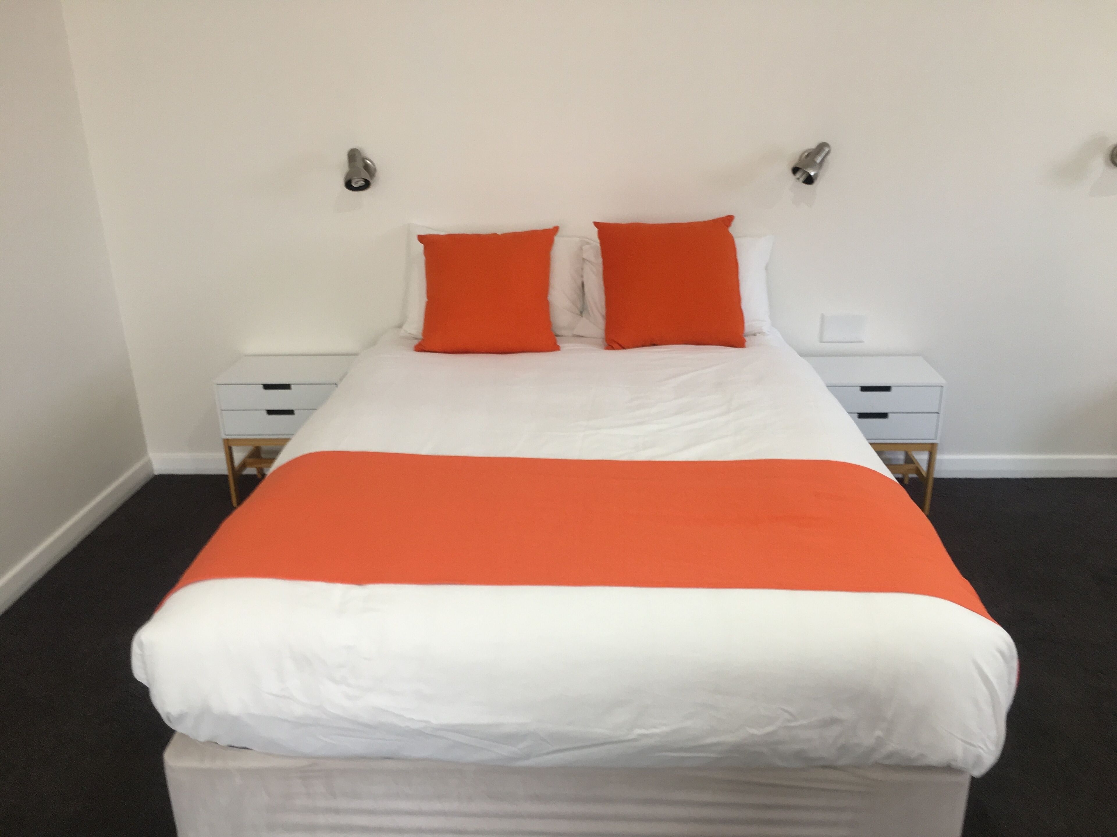 A neatly made bed with white and orange bedding, two bedside tables with drawers, and a plain white wall with two wall-mounted light fixtures.