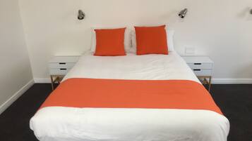 A neatly made bed with white and orange bedding, two bedside tables with drawers, and a plain white wall with two wall-mounted light fixtures.