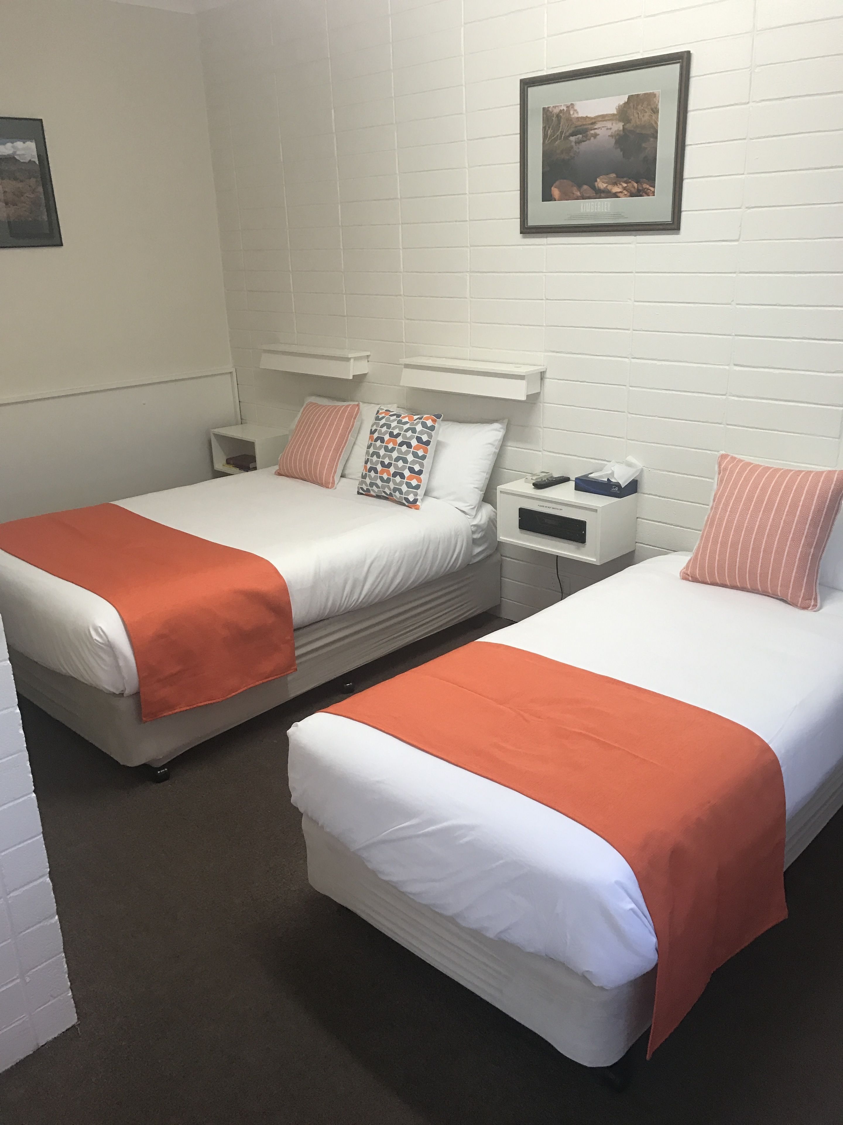 A hotel room with two single beds, each with an orange blanket, white bedding, and a nightstand with a remote control and tissues.