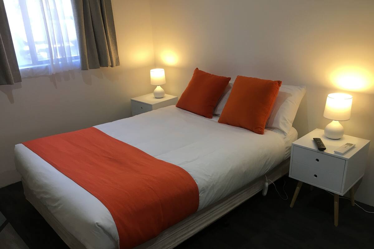A neatly made bed with an orange throw blanket, two white pillows, and two orange pillows. There are two bedside tables with lamps, one on each side of the bed.