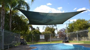 Una piscina con zona sombreada, rodeada por una valla metálica, bancos y un cartel que indica que es una piscina infantil.