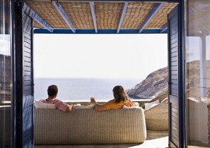 A room with a large window overlooking a coastal view, featuring a wicker sofa and a blue ceiling. - Aegea Blue Cycladic Resort (Andros)