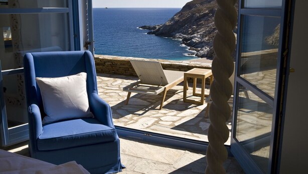A room with a view of the ocean, a blue chair, a small table, and a stone floor.