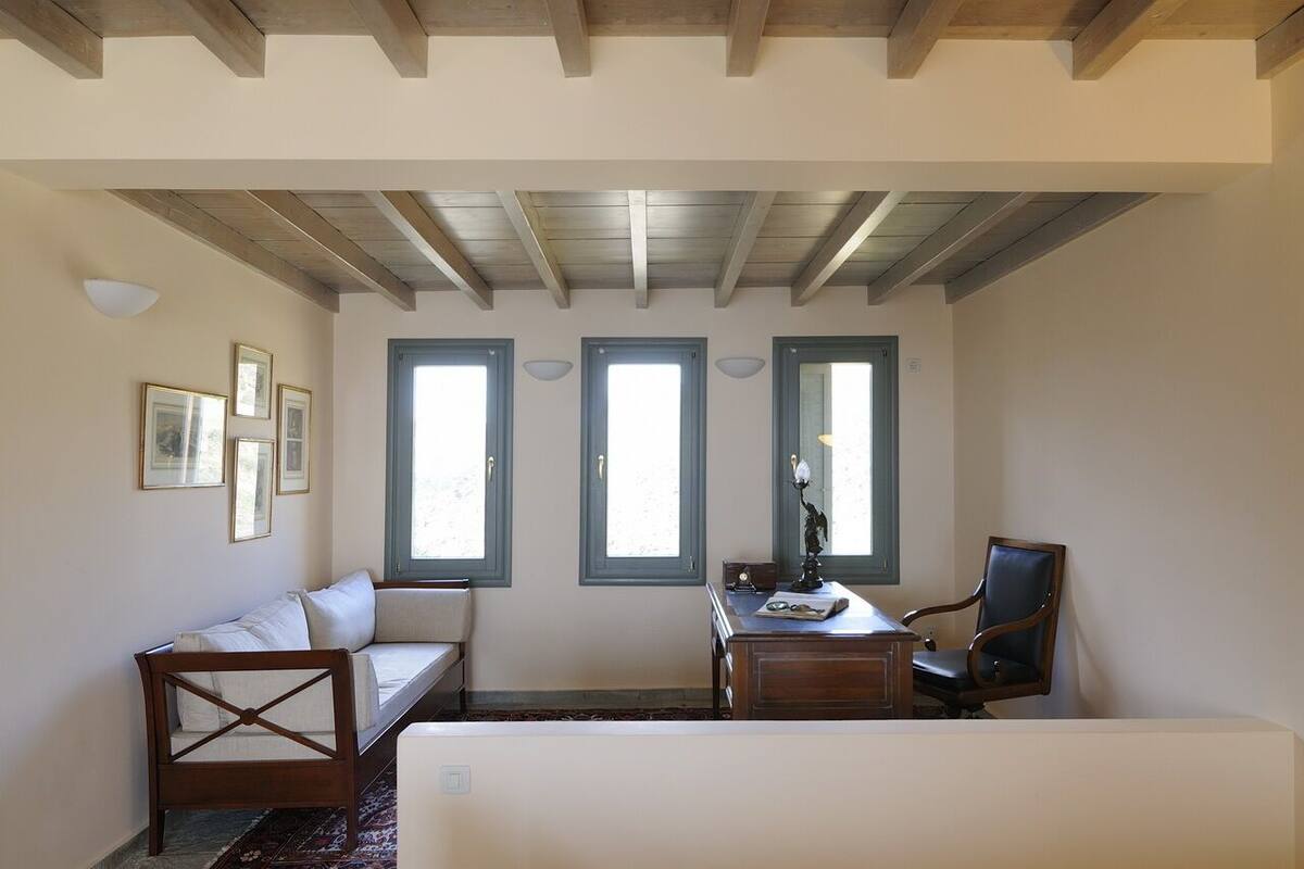 Une pièce avec un plafond en bois, un canapé, un bureau avec une chaise et trois fenêtres équipées de volets.