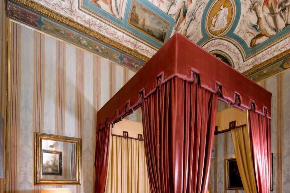 A four-poster canopy bed with red bedding, a round table with a red lamp, and a patterned rug.