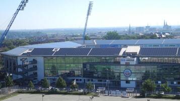 A modern sports stadium with solar panels on the roof, surrounded by a parking lot and a cityscape in the background.