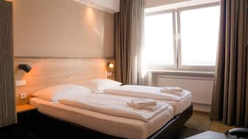 Habitació d'hotel amb dos llits, un capçal de llit de fusta, una tauleta de nit, una làmpada i una finestra amb cortines.