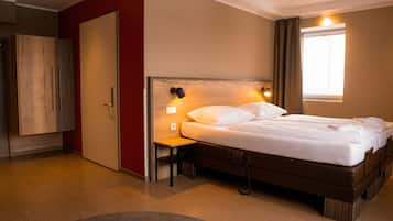 Quarto de hotel moderno com uma cama grande, mesa de cabeceira, guarda-roupa e janela com cortinas.