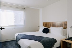 A hotel room with a large bed, a wooden headboard, a bedside table with a lamp, and a window with blinds.