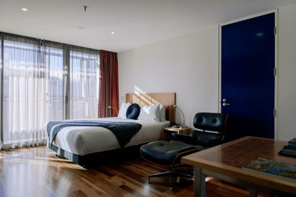 A hotel room with a large bed, a desk, a chair, and a dark-colored door.