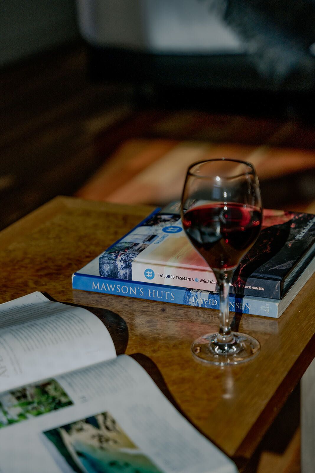 A glass of red wine on a wooden table with a book titled "Mawson's Huts" and another book by David Nissen.