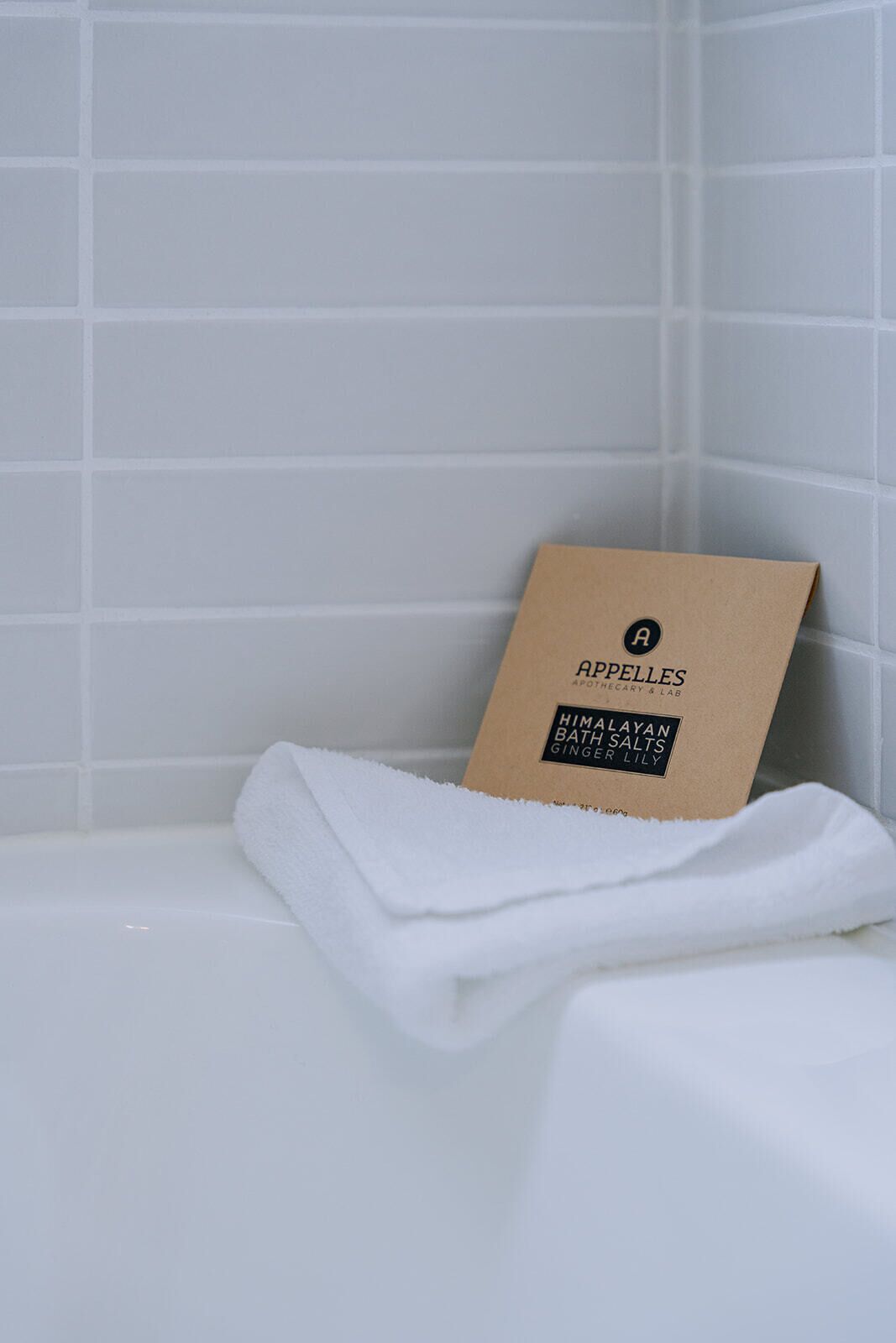 A bathroom with white tiles, a white towel, and a brown box labeled "ARPELE'S APOTHECARY & LAB. HIMALAYAN BATH SALTS GINGER LILY."