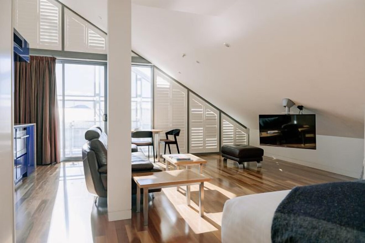 A modern loft-style bedroom with a sloped ceiling, a large window, a flat-screen TV, and a seating area with a table and chairs.