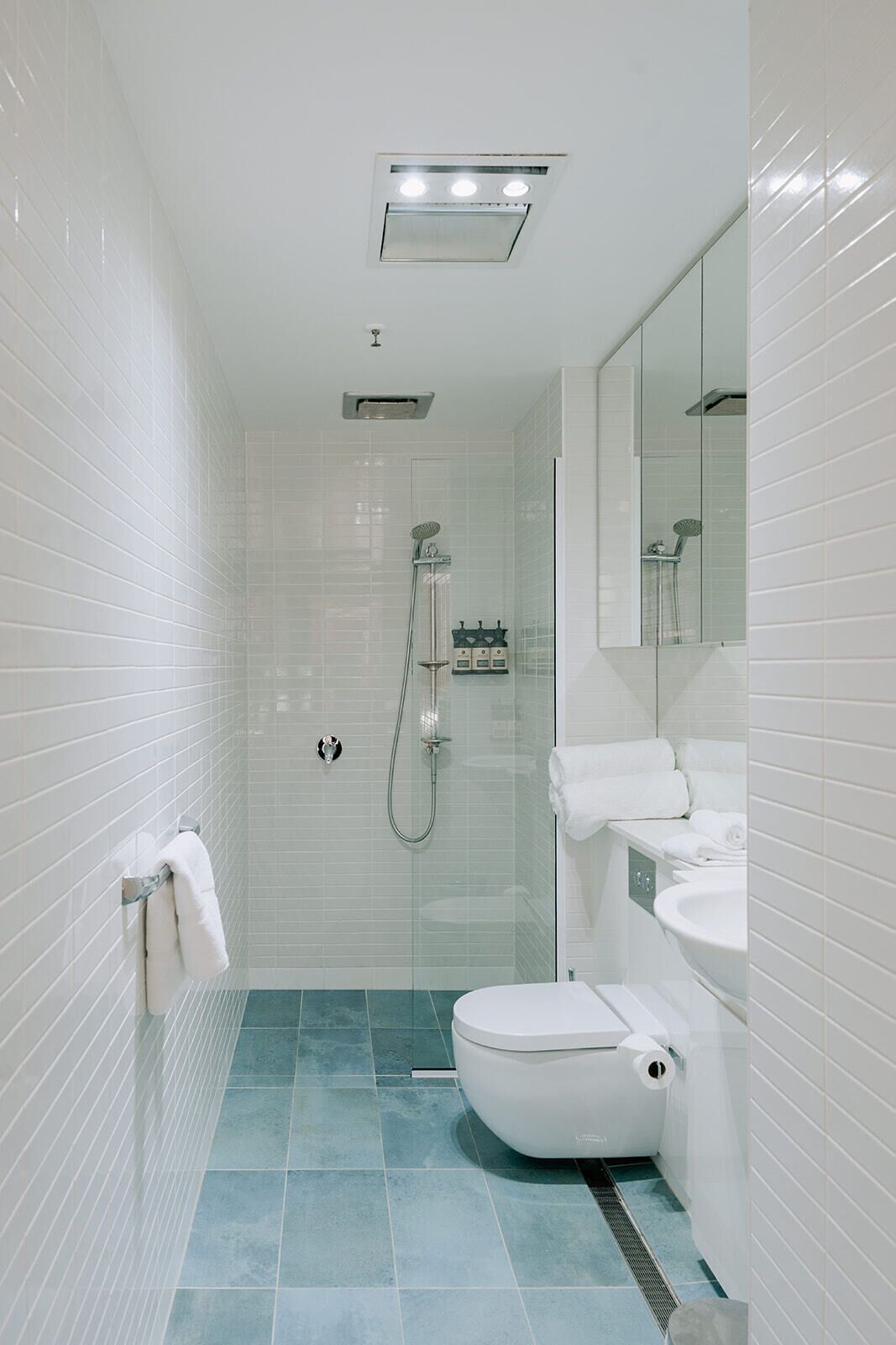 Um banheiro moderno com paredes em azulejo branco, box de vidro, vaso sanitário e pia com espelho.