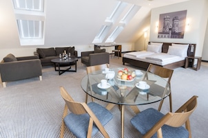 Habitación de hotel moderna con cama altillo, un sofá, una mesa de comedor de vidrio y vistas a un paisaje urbano a través de tragaluces.