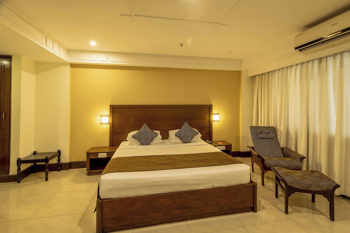 Una habitación de hotel con una cama grande, dos mesitas de noche, una silla, un buró y un equipo de aire acondicionado.