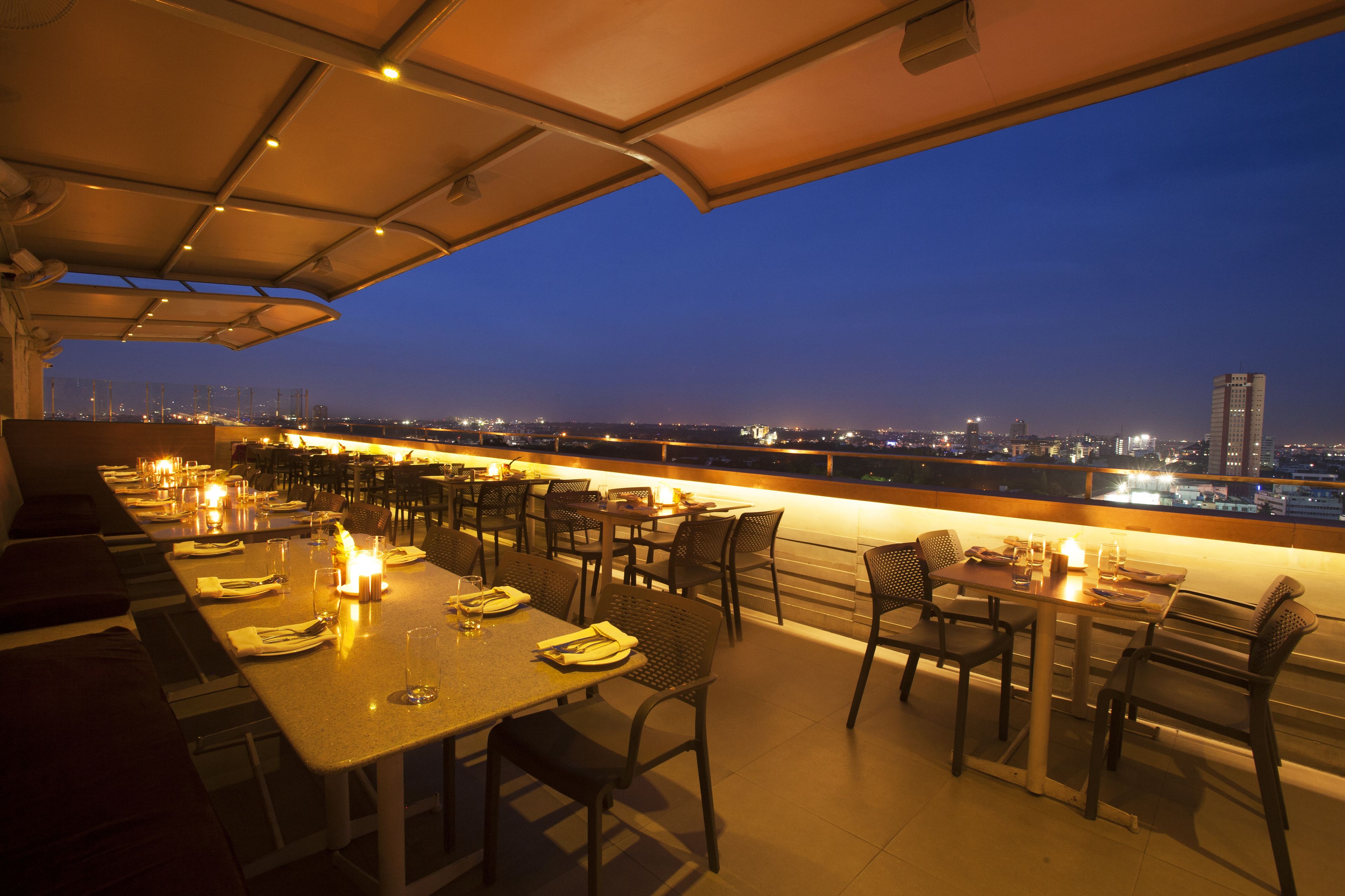 A rooftop restaurant with tables and chairs, a city view, and a railing.
