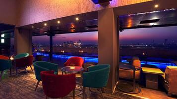 Un salon moderne avec des chaises et des tables aux couleurs vives, de grandes fenêtres offrant une vue sur la ville et un espace piscine éclairé de lumières bleues.