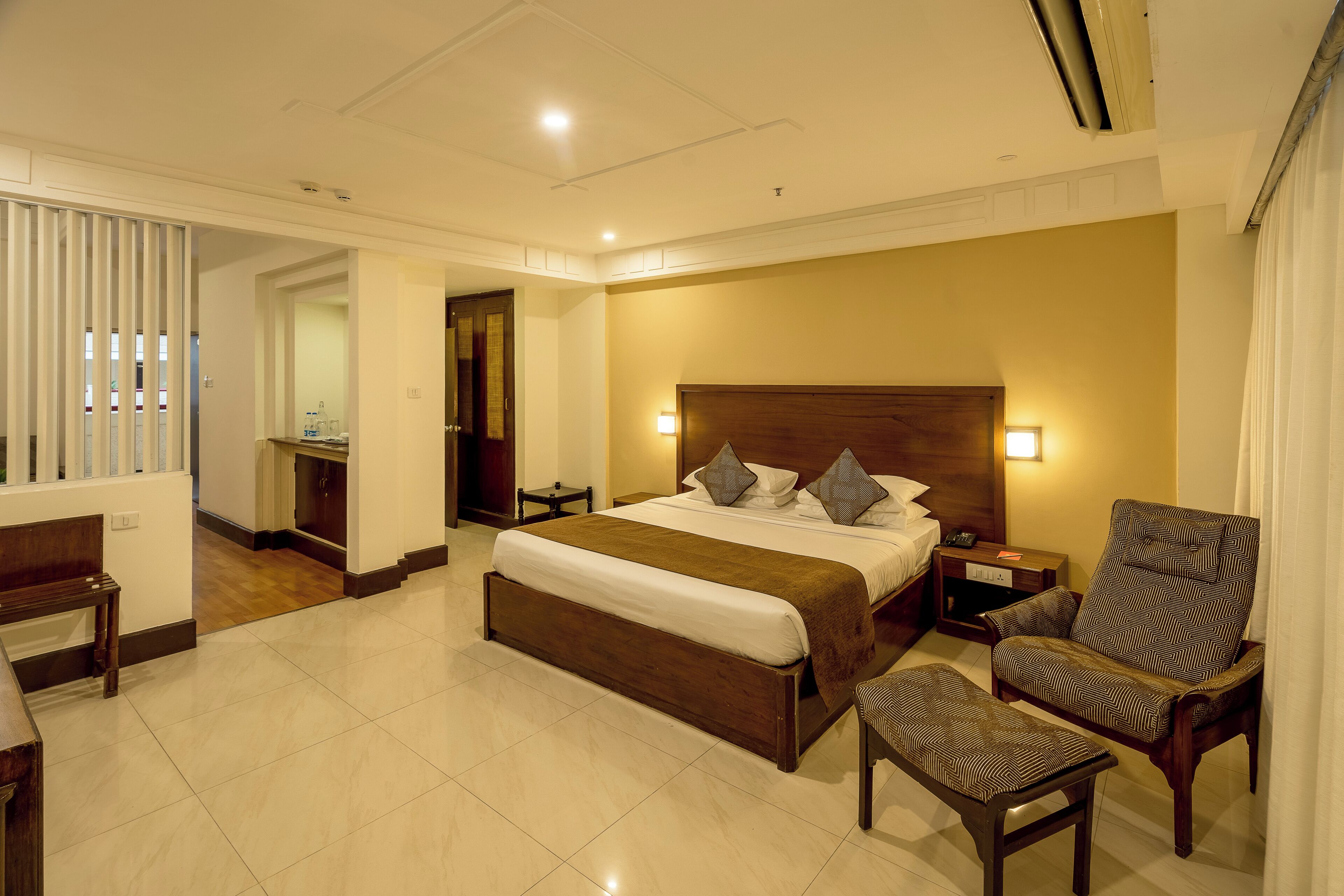 A hotel room with a large bed, a wooden headboard, a nightstand, a chair, and a bench.