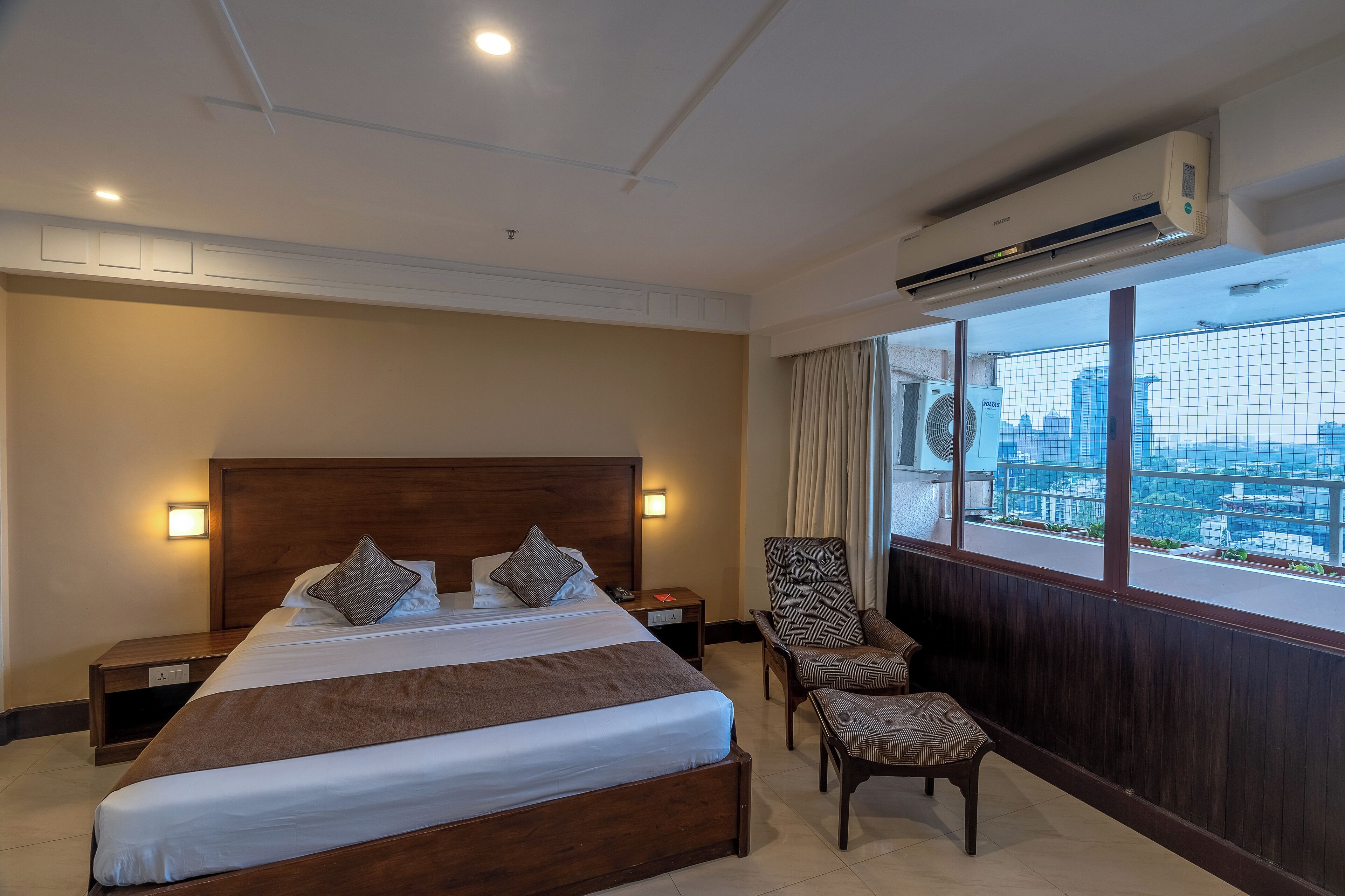 A hotel room with a large bed, a wooden headboard, bedside tables, a chair, and a view of a cityscape through large windows.