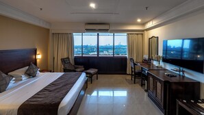 Een hotelkamer met een groot bed, een bureau met een televisie, een stoel en uitzicht op de stad door grote ramen.