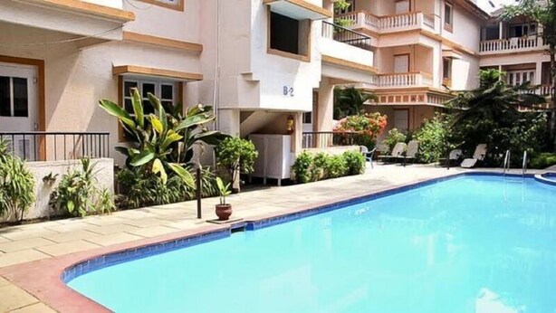 A swimming pool with a tiled edge, surrounded by potted plants and a building with balconies. - Perola Do Mar (Candolim)