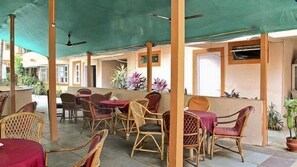 Relax in a shaded outdoor space with wicker chairs and small tables, surrounded by lush greenery and potted plants.
