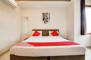 A hotel room with a bed, red pillows, a white bedspread, a framed picture on the wall, and a window with curtains.