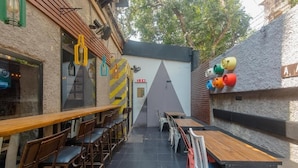 An outdoor dining area with wooden tables and chairs, a triangular wall design, and colorful cylindrical objects on the wall. - Hotel Jayshree (Mumbai)