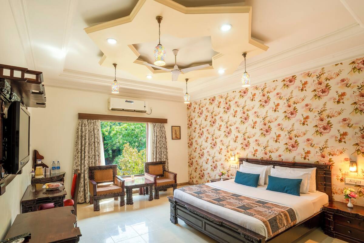 A bedroom with a large bed, floral wallpaper, a ceiling fan, and a view of greenery outside.