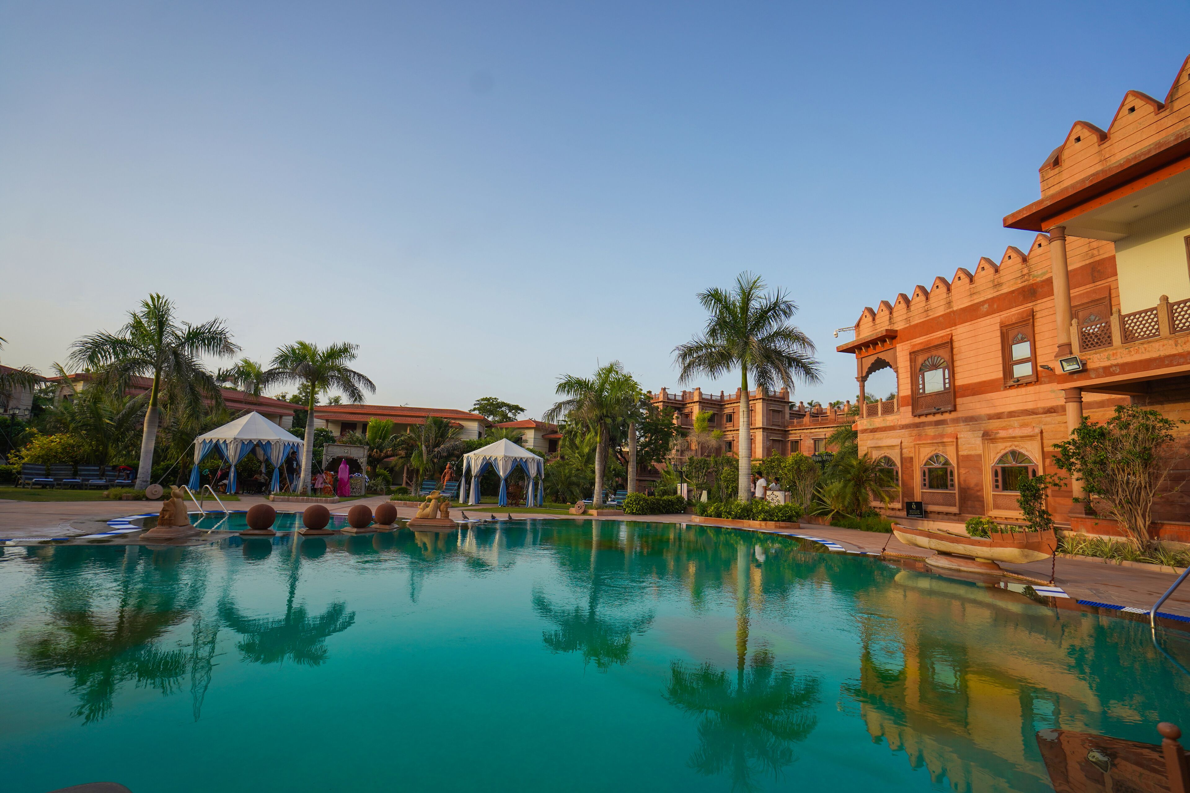 A pool area with a clear blue pool, palm trees, and a building with traditional architecture.
