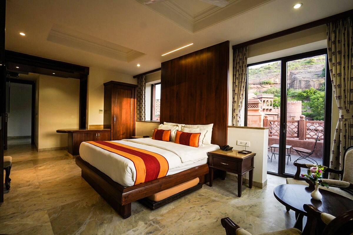 A spacious hotel room with a large bed, a small desk, a chair, and a balcony with a view of the city.