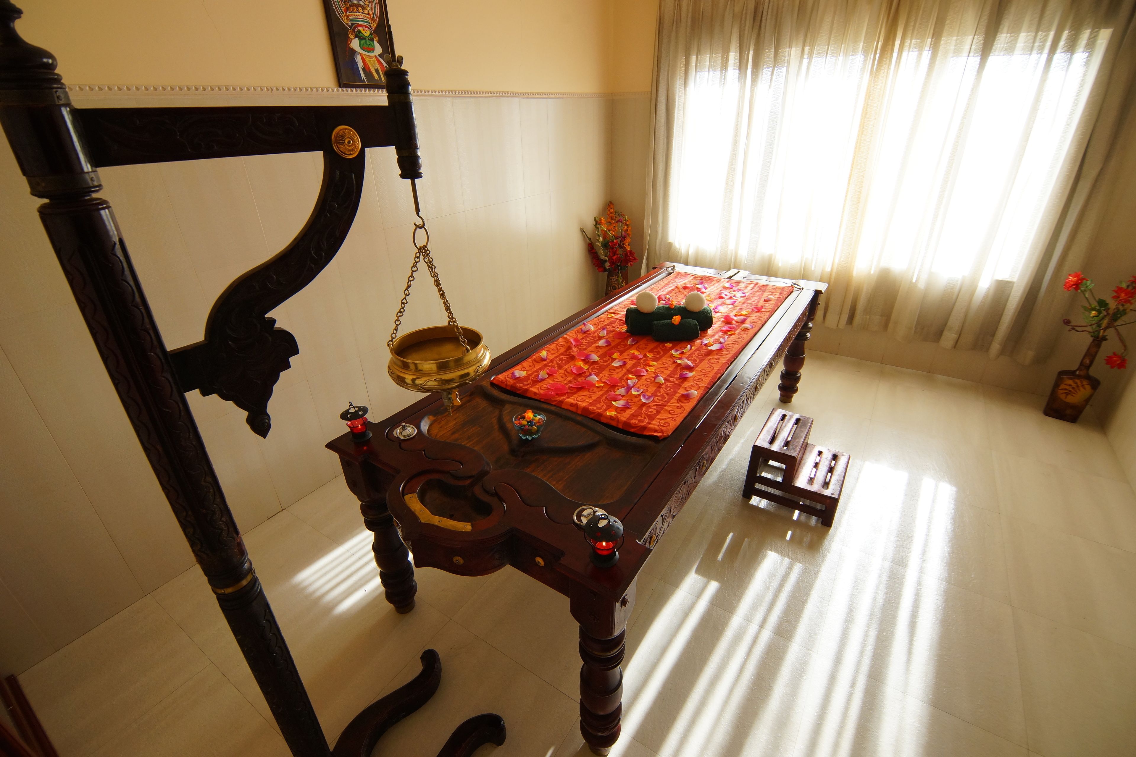A traditional room with a wooden table, a brass oil lamp, and a bed with a red bedspread.