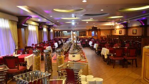 A banquet hall with long tables, red chairs, and a central buffet area. - Joys Palace (Thrissur)