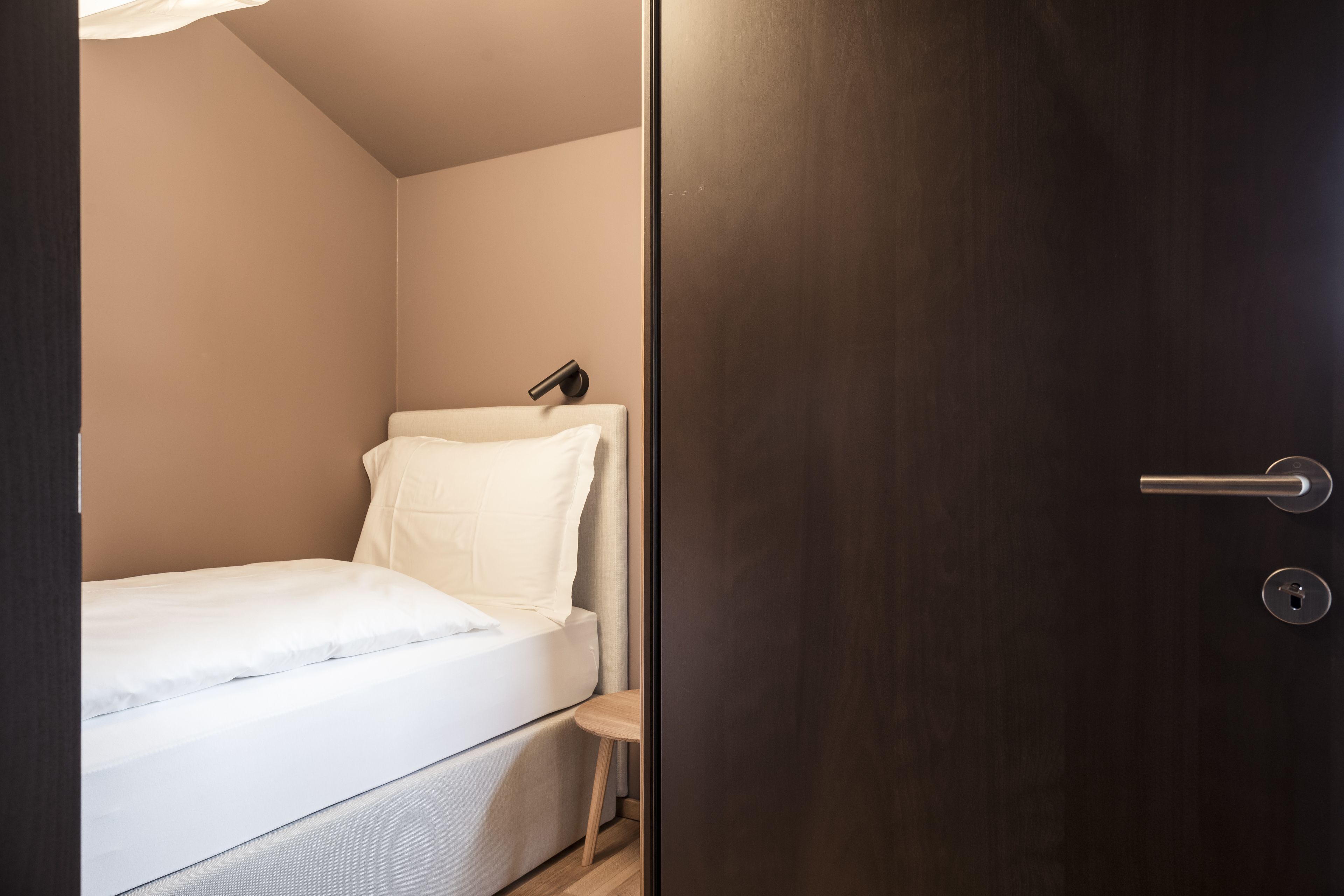 A small, neatly made bed in a compact room with a dark wooden door and a small table beside it.