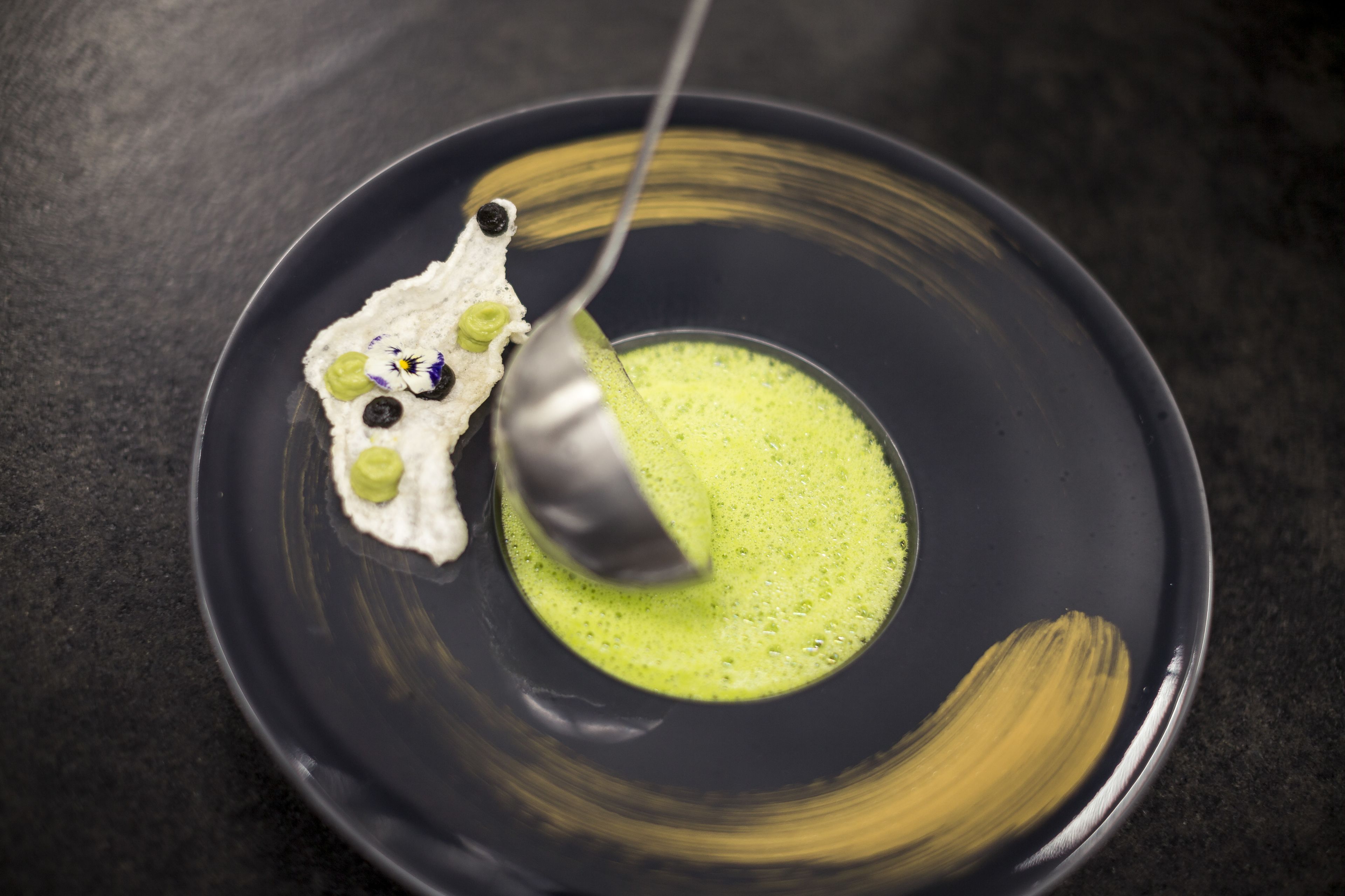 Une cuillerée de sauce verte sur une assiette noire, accompagnée d’une crème blanche et d’éléments décoratifs.