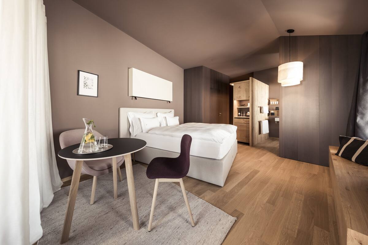 A modern hotel room with a bed, a small table with a chair, a bedside table with a lamp, and a view into the kitchen area.