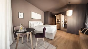Ein modernes Hotelzimmer mit einem Bett, einem kleinen Tisch mit Stuhl, einem Nachttisch mit Lampe und Blick in die Küchenzeile.