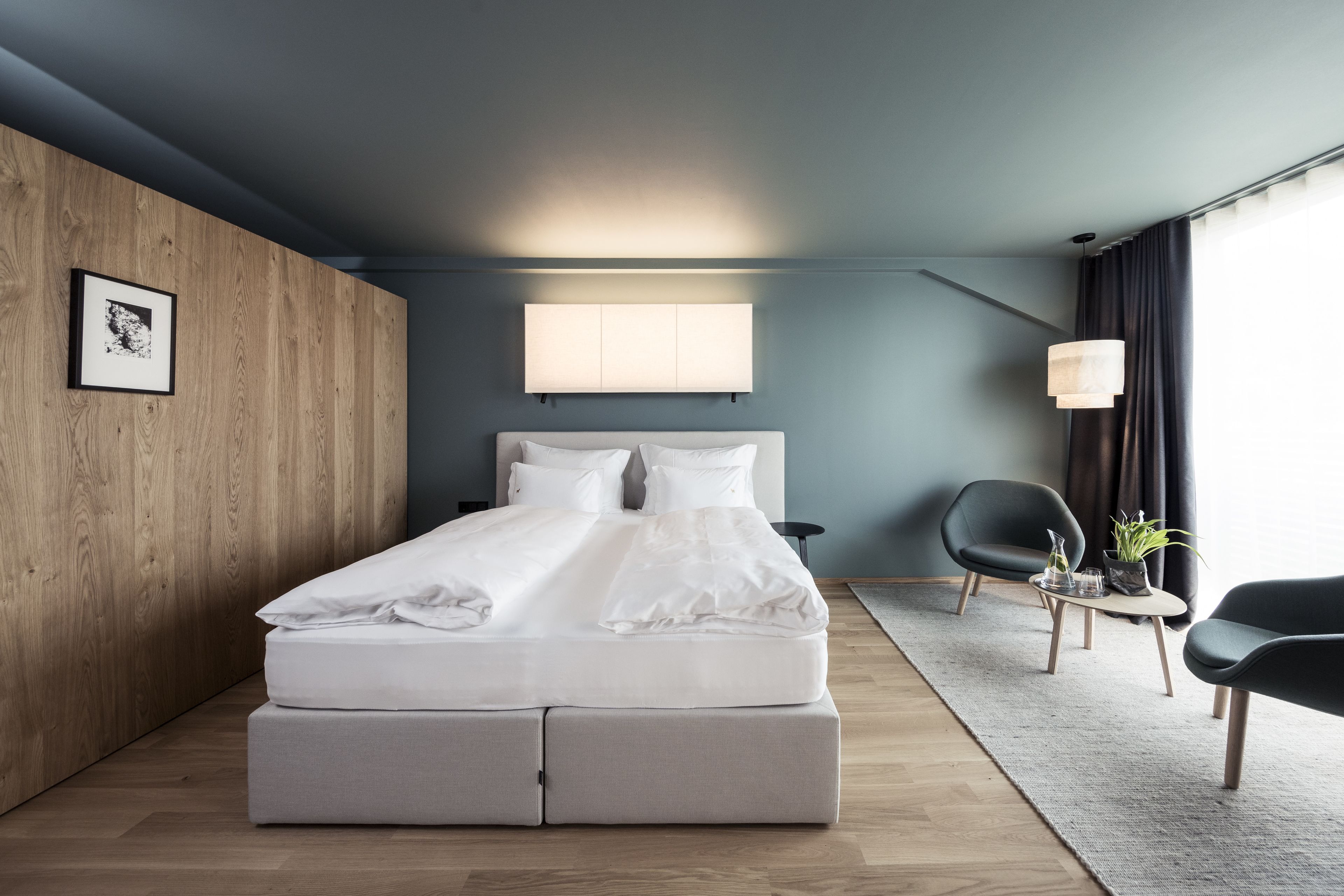 A modern bedroom with a bed, a wooden accent wall, a bedside table, a chair, and a window with curtains.