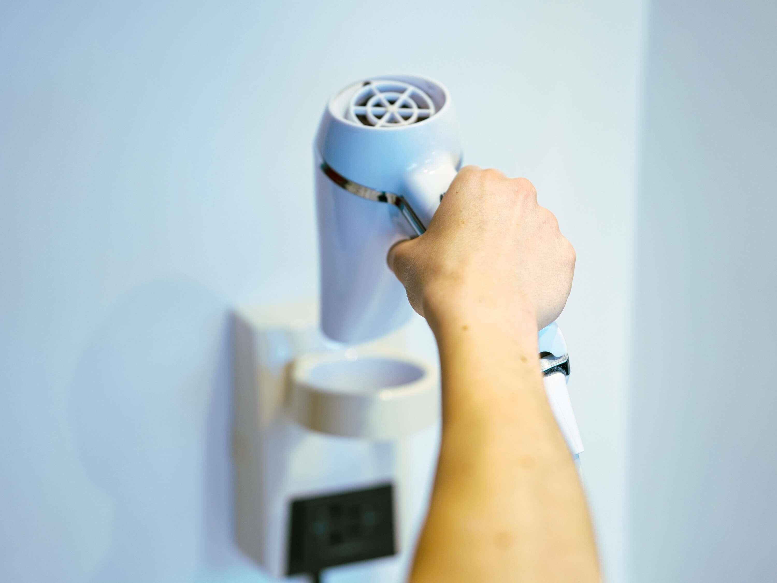 A hand holding a white device with a circular pattern on top, possibly a hairdryer, against a plain wall.