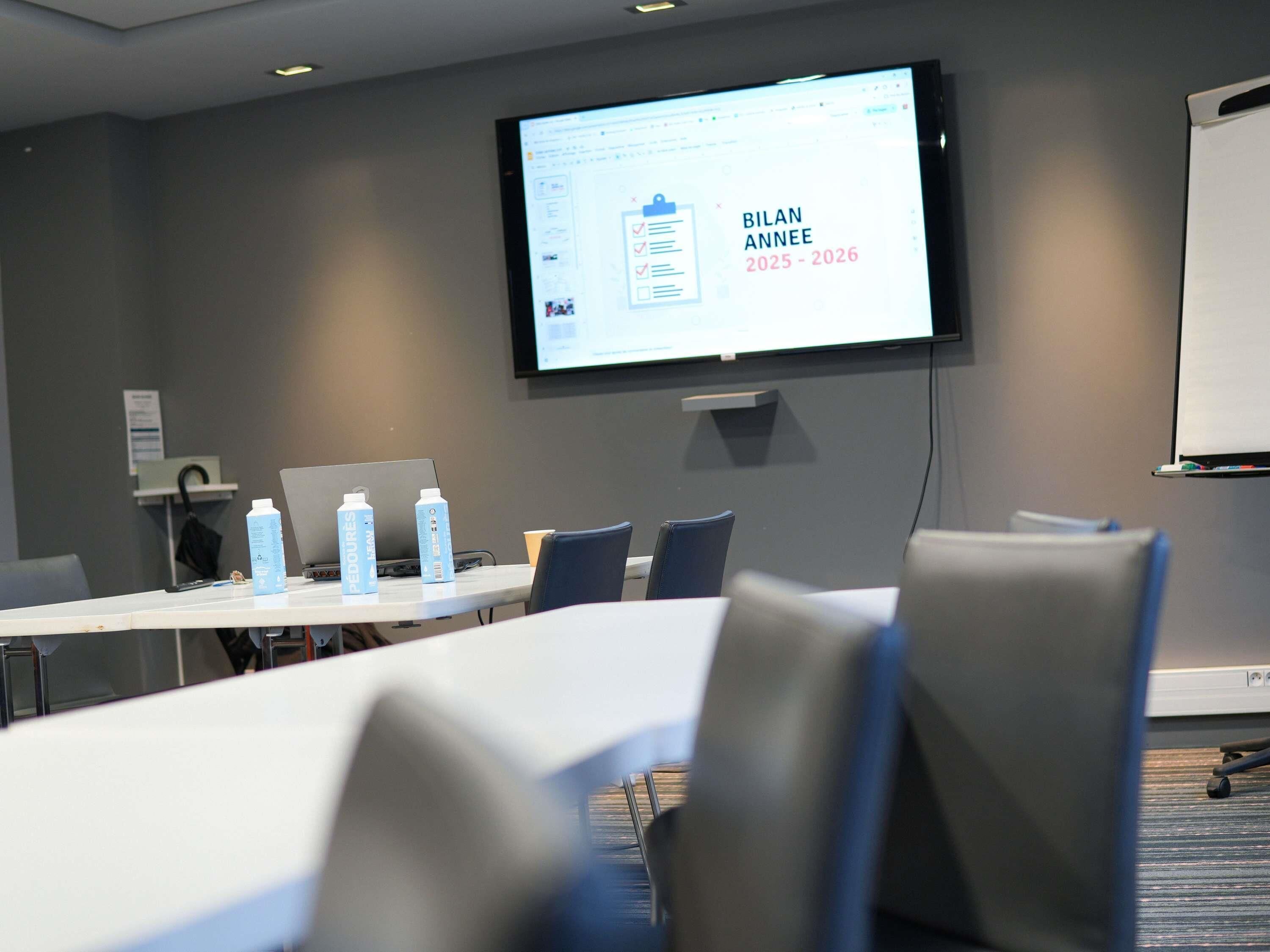 Sala de conferência com uma tela grande exibindo uma apresentação, um quadro branco e várias cadeiras.